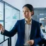 Femme gestionnaire de projet en costume bleu, analysant des graphiques et un tableau de bord sur de grands écrans interactifs dans un bureau.