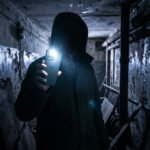 Une personne en sweat à capuche tient un smartphone éclairé dans un couloir sombre et délabré.